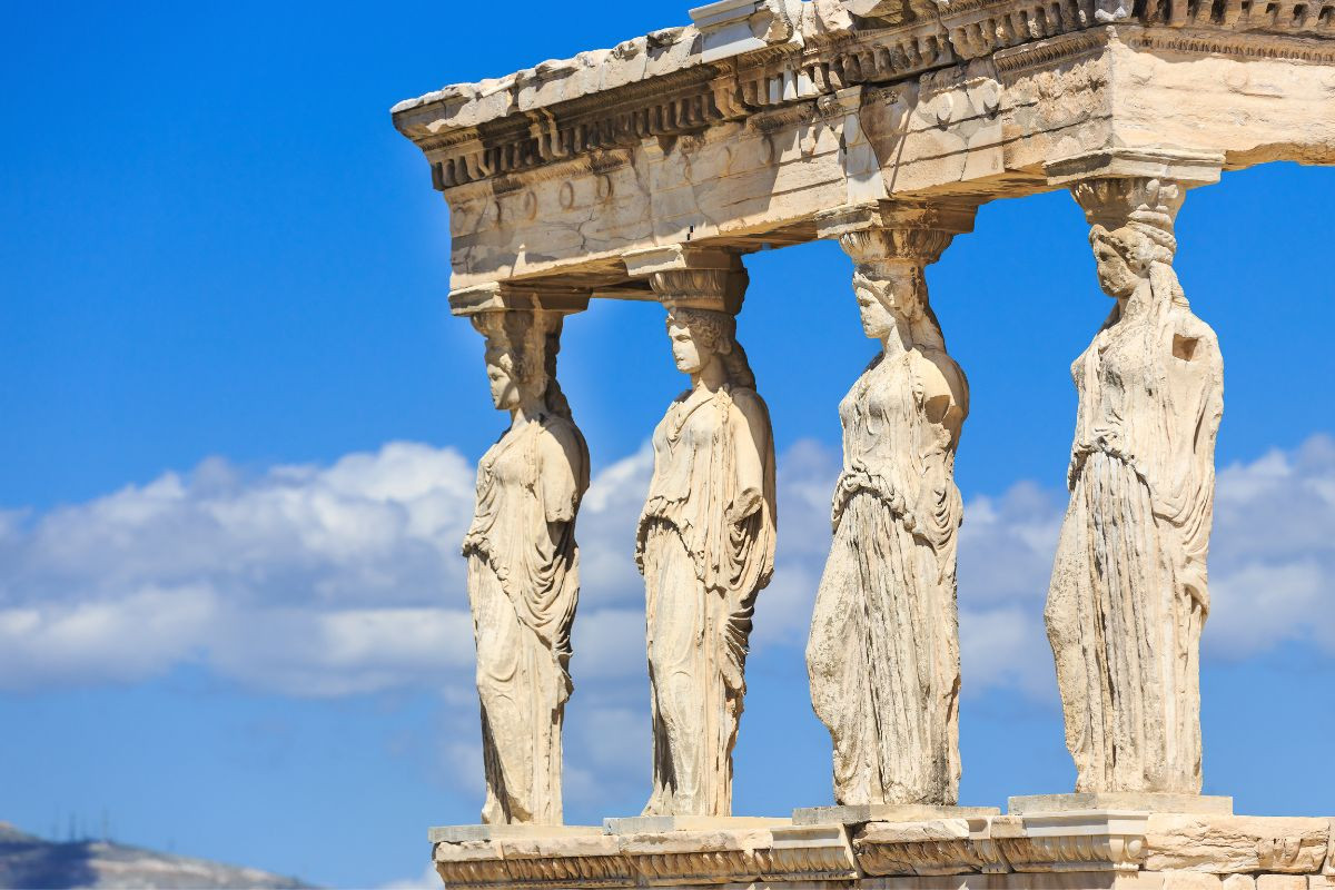 The image shows the Caryatids, a set of six ancient Greek female statues, which serve as architectural supports on the Erechtheion temple in Athens, Greece. The statues are aligned in a row, each draped in traditional attire, against a backdrop of a clear blue sky.