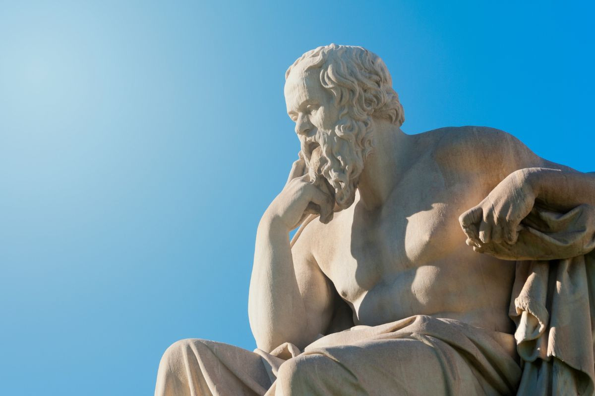 The image shows a statue of the ancient Greek philosopher Socrates in a thoughtful pose, with his hand resting on his chin. The statue is set against a clear blue sky.